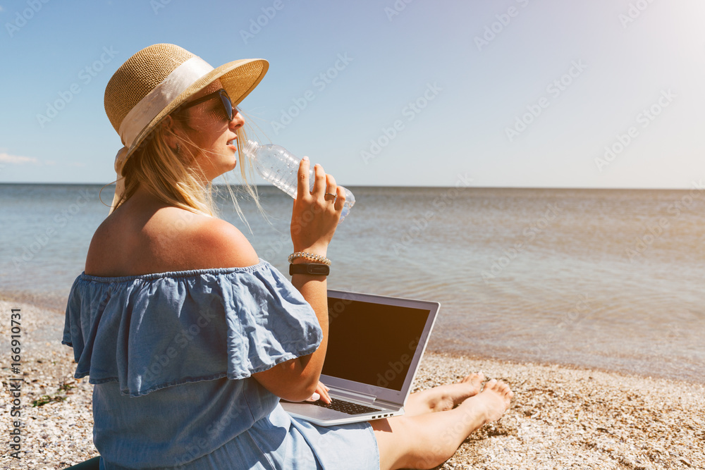 Fototapeta premium Young beautiful woman sitting with laptop on the beach near the sea and drinking water