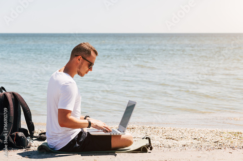Young man in white T-shirt sits on the beach and working with laptop