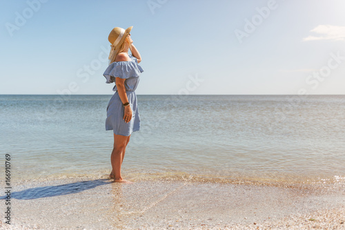 Young beautiful woman in blue dress walks along the beach at sea