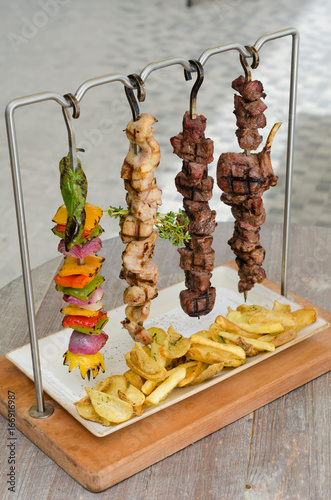 Cooked Chicken, beef and vegetable kebab skewers hanging from a rack, served with fries, and ready to eat.