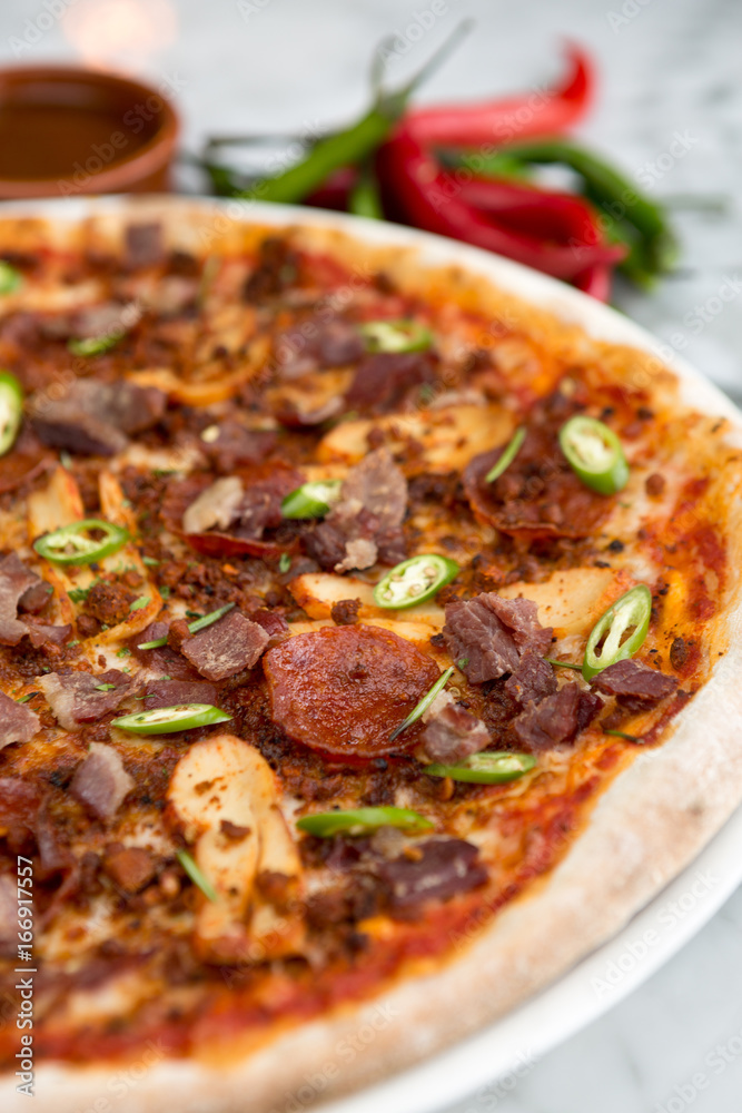 Spicy pepperoni, chicken and bacon pizza with jalapeño slices and chilli peppers in the background Spicy pepperoni, chicken and bacon pizza with jalapeño slices and chilli peppers in the background