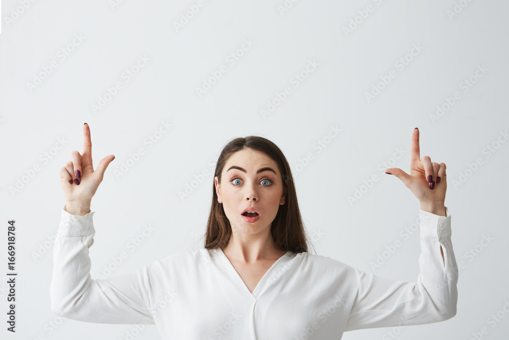 Surprised young beautiful brunette businesswoman smiling looking at camera pointing fingers up over white background. Surprised young beautiful brunette businesswoman smiling looking at camera pointing fingers up over white background.