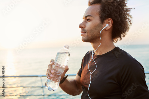 Fotografija Outdoor shot of stylish dark-skinned male athlete drinking water out of plastic bottle after cardio workout