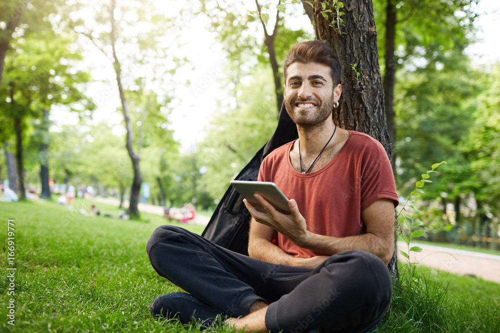 Stylish bearded man with appealing dark eyes smiling into camera having pleased expression sitting cross-legged on grass, reading from tablet. Casually dressed student studying outdoors, using tablet. Stylish bearded man with appealing dark eyes smiling into camera having pleased expression sitting cross-legged on grass, reading from tablet. Casually dressed student studying outdoors, using tablet.