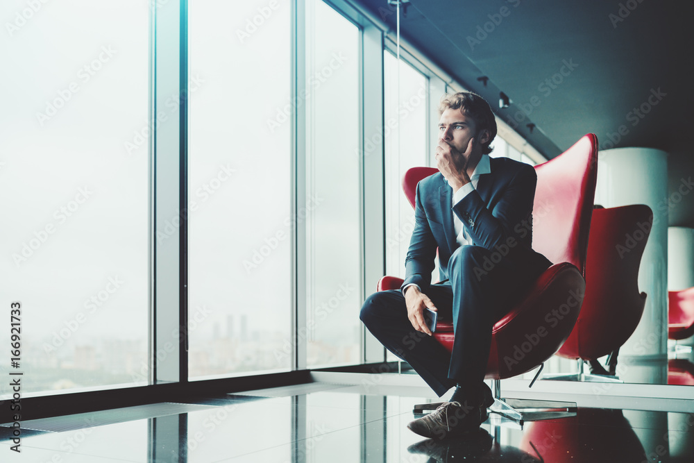Adult pensive and anxious businessman in blue formal suit is sitting on red armchair in luxury office interior near window and recapitulating recent work meeting with copy space zone for advertising - obrazy, fototapety, plakaty 