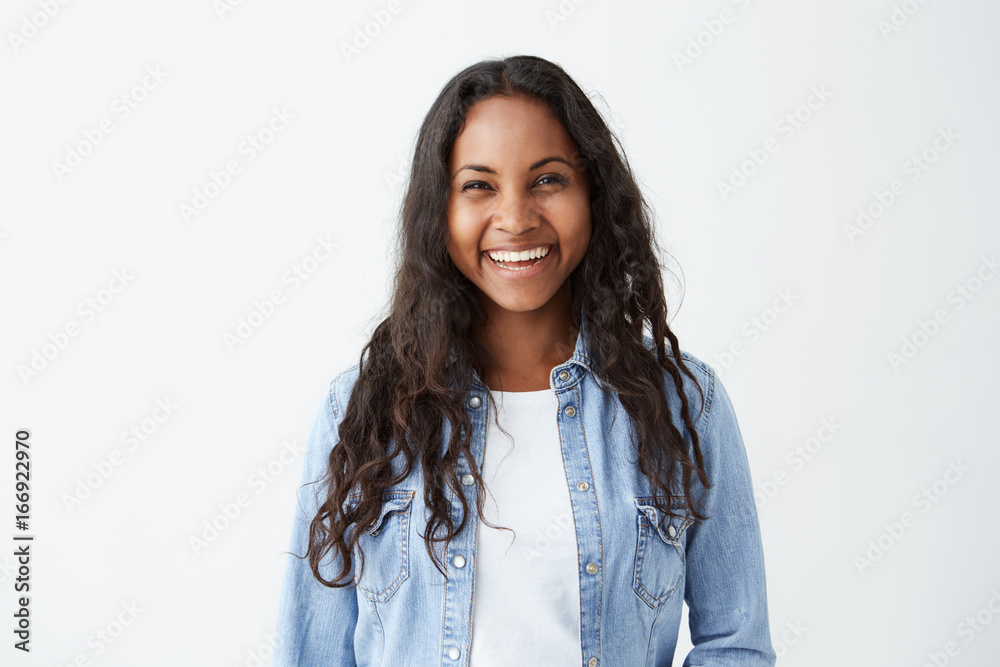 Beautiful dark-skinned female with long brunette hair and broad happy smile wearing denim shirt enjoying good positive news concerning her promotion at work, posing isolated against white blank wall Beautiful dark-skinned female with long brunette hair and broad happy smile wearing denim shirt enjoying good positive news concerning her promotion at work, posing isolated against white blank wall