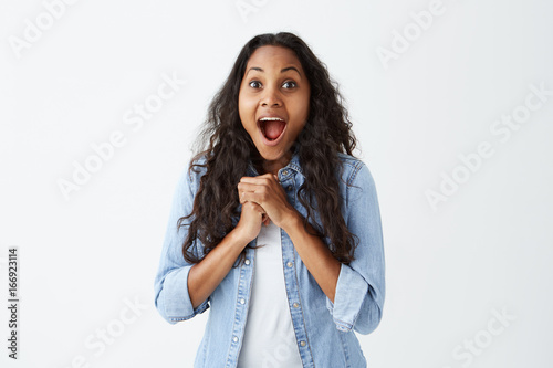 Fotografi Beautiful dark-skinned woman in casual clothes with open mouth celebrating her victory keeping clenched fists together having excited and satisfactied expression