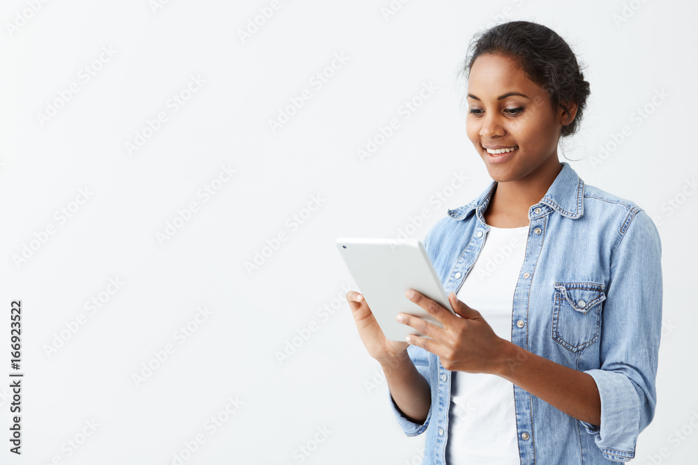 Cheerful young dark-skinned female student with cute smile standing on white background, using tablet, checking newsfeed on her social network accounts. Pretty afro-american girl surfing internet on Cheerful young dark-skinned female student with cute smile standing on white background, using tablet, checking newsfeed on her social network accounts. Pretty afro-american girl surfing internet on