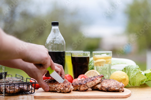 Male hand cut chicken breast grilled on a wooden Board. Food photography chicken chili steak on the background with vegetables and black soda and lemonade. Food and summer drinks for a picnic. photo