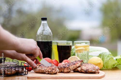 Male hand cut chicken breast grilled on a wooden Board. Food photography chicken chili steak on the background with vegetables and black soda and lemonade. Food and summer drinks for a picnic. photo