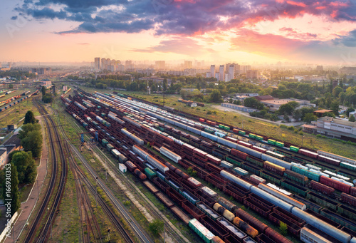 Aerial view of colorful freight trains. Railway station. Cargo trains. Wagons with goods on railroad. Heavy industry. Industrial scene with trains, city buildings and cloudy sky at sunset. Top view 