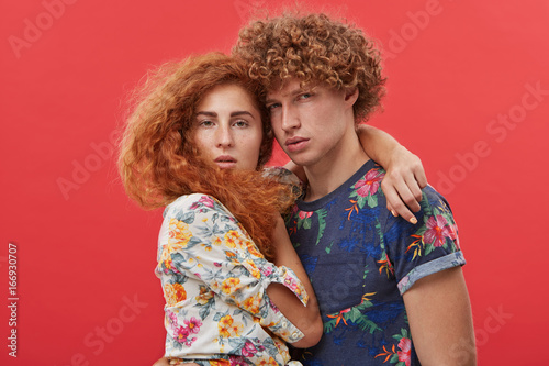 Style, fashion, beauty and glamour. Headshot of stylish gorgeous ginger female model with long bushy hair embracing confident attractive red-haired male, both staring at camera with serious looks