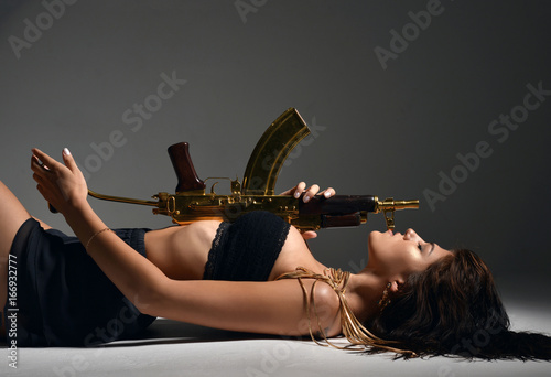 sexy brunette woman lying with fashion gold automatic rifle gun on gray background