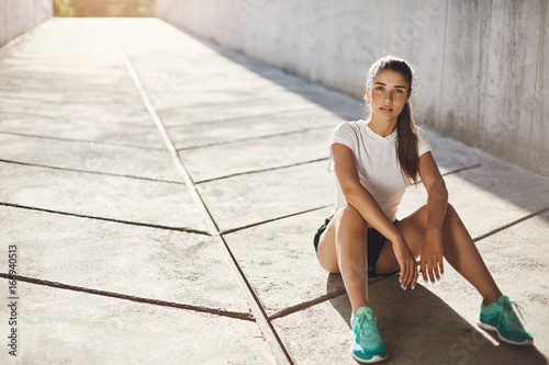 Fototapeta Naklejka Na Ścianę i Meble -  Young lady runner sitting on a pavement getting ready for a long run looking at camera. Urban sport concept.