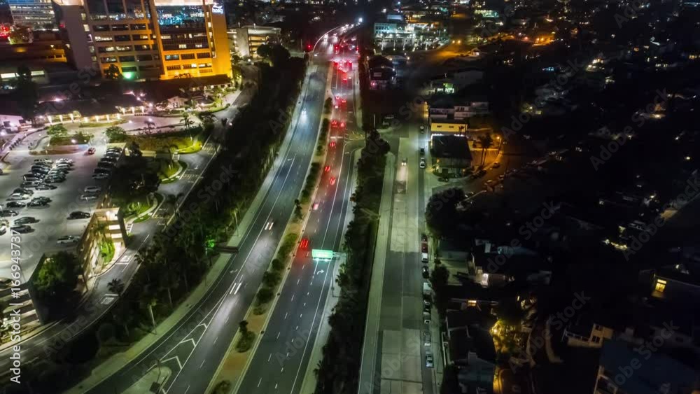 Aerial motion timelapse or hyper lapse at night over a traffic circle onramp and up a street showing streaks of car lights, city buildings and parking lots, indicative of the fast paced city lifestyle