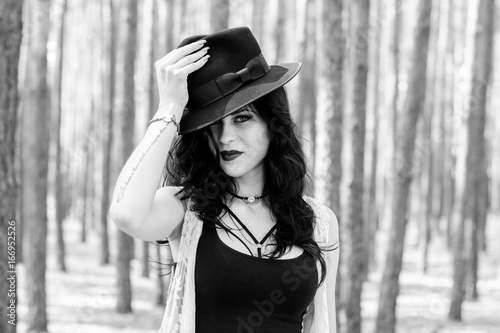  Enigmatic curly brunette woman with gothic make up and black fedora hat, white lace cardigan, in boho style walk in forest and thinking about life, near forest at autumn, stylish woman on nature