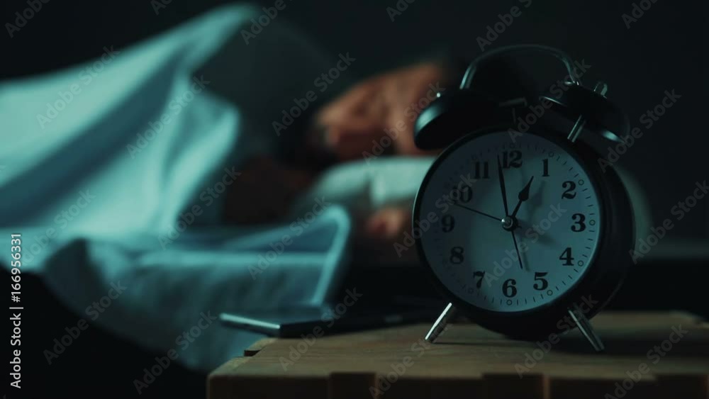 Adult caucasian man talking on mobile phone late at night while lying in bed in bedroom, selective focus on alarm clock on bedside table