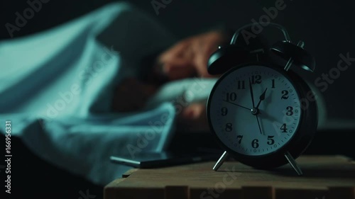 Adult caucasian man talking on mobile phone late at night while lying in bed in bedroom, selective focus on alarm clock on bedside table