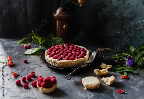 raspberry tart with fresh beries on old black pan with coffee grinder, spoon. flowers and crushed tart on a grey background