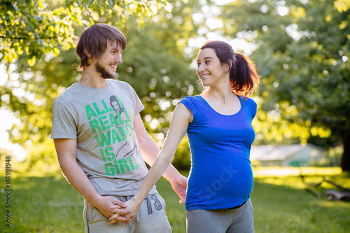 Bright picture of hipster man and pregnant woman in sports wear are holding by hands and looking at each other in beautiful summer day in green park.