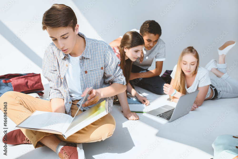 teenage boy reading book while multicultural friends using laptop isolated on white teenage boy reading book while multicultural friends using laptop isolated on white