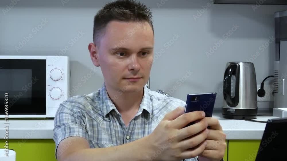 Handsome Young Businessman Before Working Day with Smartphone Drinking Coffee. Attractive Man at Home Using Smartphone in Kitchen Typing Text And Sending Message on Social Media at Morning Handsome Young Businessman Before Working Day with Smartphone Drinking Coffee. Attractive Man at Home Using Smartphone in Kitchen Typing Text And Sending Message on Social Media at Morning