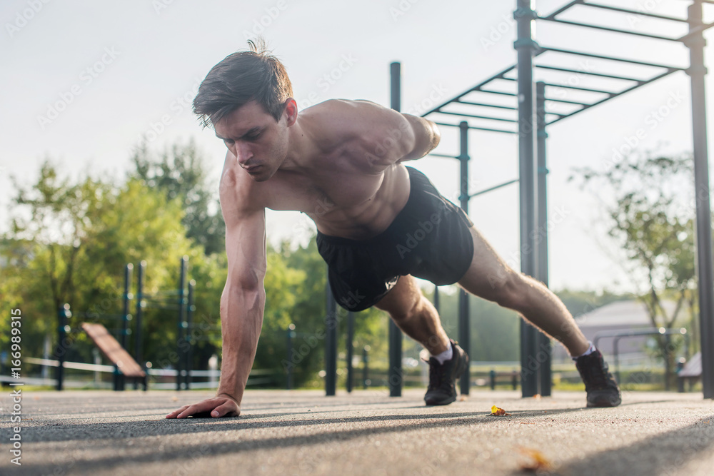 Athlete young man doing one-arm push-up exercise working out his upper body muscles outside in summer.