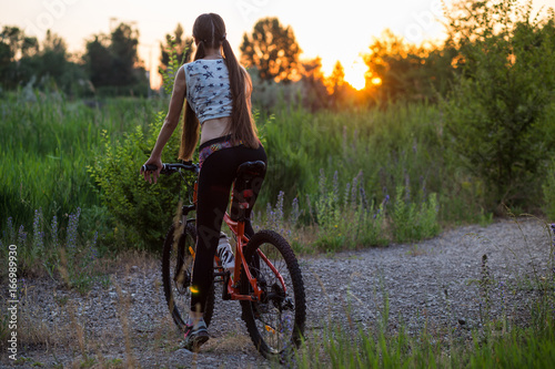 Attractive sporty girl with long hair on a bicycle drinking water from the sport bottle on sunset