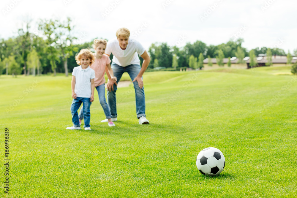 happy father with daughter and son looking at soccer ball while standing on grass