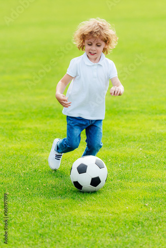 cute happy little boy playing soccer on green grass at park