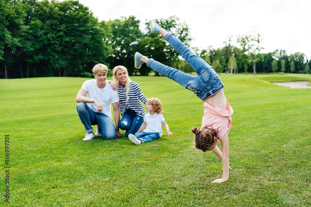 Obraz premium happy parents with son looking at cute little daughter performing handstand at park