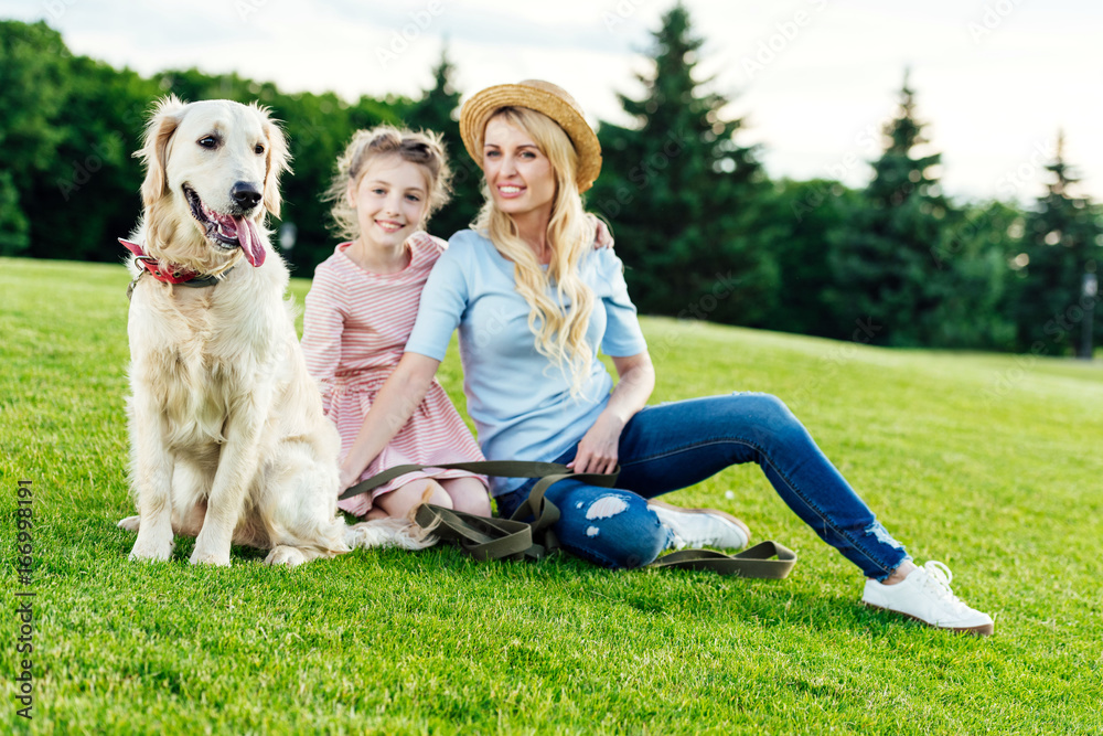 happy mother and daughter with dog sitting on green lawn at park happy mother and daughter with dog sitting on green lawn at park