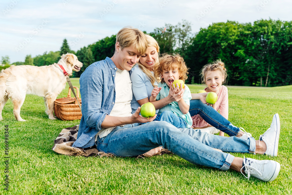 Fototapeta premium happy young family eating apples while resting with pet at picnic