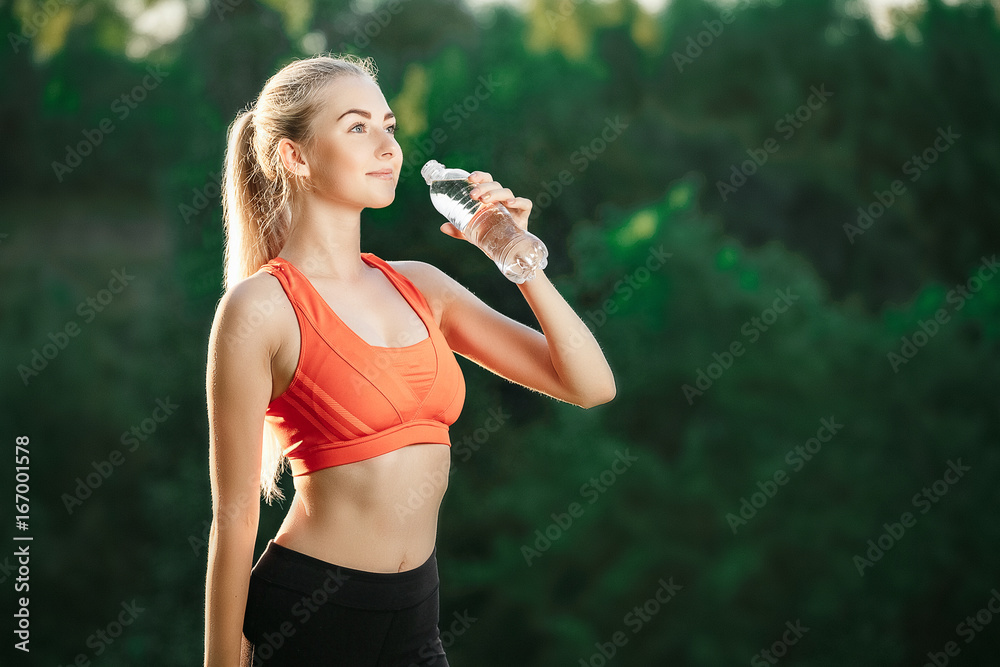 A young blonde in a red top and black pants holds a bottle of water in the hands. Concept of healthy eating and sport. A young blonde in a red top and black pants holds a bottle of water in the hands. Concept of healthy eating and sport.