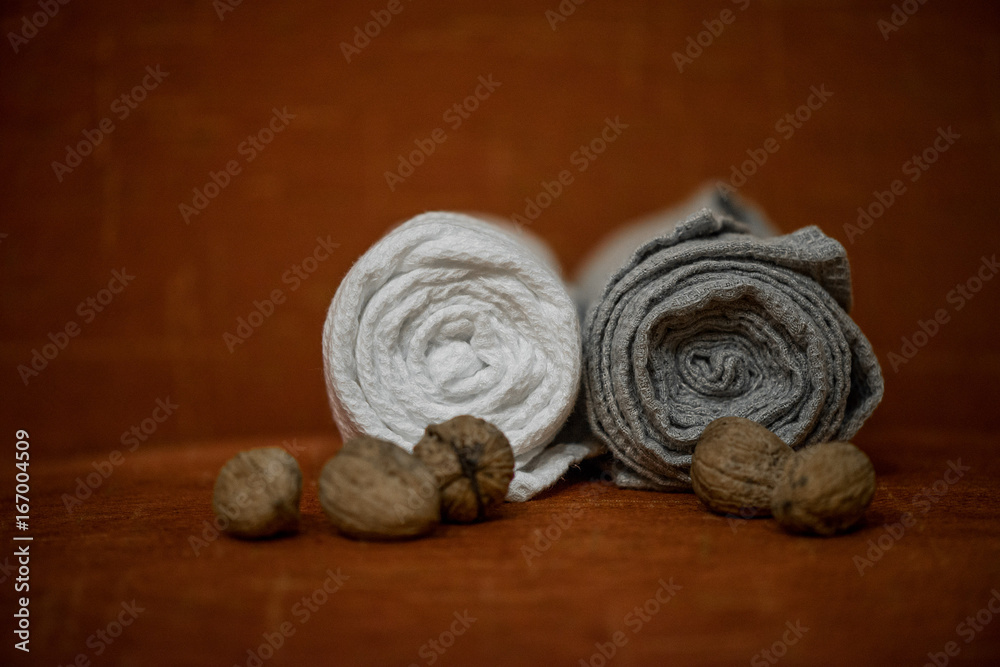 Macro shot of rolled, folded snow white and grey natural linen sauna and bath towels next to small walnuts on orange background, on massage table. Beautiful home decor, gift for newlyweds for wedding Macro shot of rolled, folded snow white and grey natural linen sauna and bath towels next to small walnuts on orange background, on massage table. Beautiful home decor, gift for newlyweds for wedding