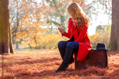 Beautiful young woman siting on vintage suitcase in a autumn park and looking at phone.