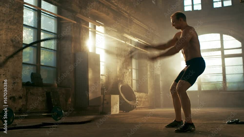 Strong Muscular Shirtless Sportsman Works out Hard with Battle Ropes as Part of His Cross Fitness Training Routine. He's Covered in Sweat and Exercises in a Abandoned Factory Remodeled into Gym. Strong Muscular Shirtless Sportsman Works out Hard with Battle Ropes as Part of His Cross Fitness Training Routine. He's Covered in Sweat and Exercises in a Abandoned Factory Remodeled into Gym.