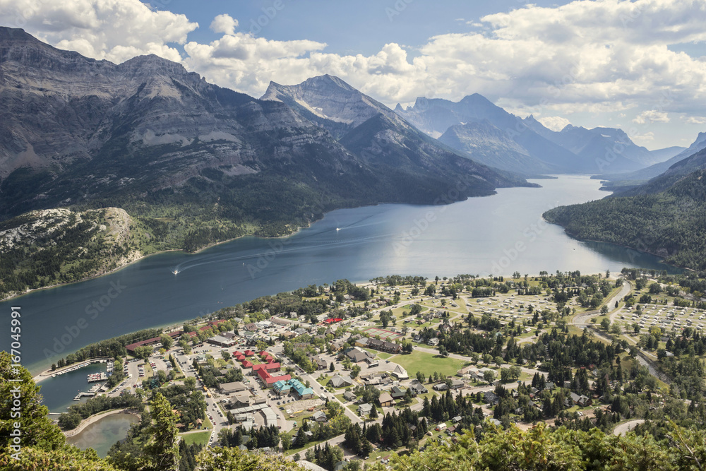 horizontal image taken from a mountain top looking down far below on to a small town of Waterton with a lake surrounded by mountains at Waterton National Park in the summer.