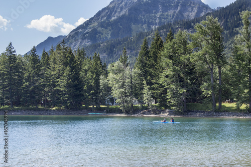 man and woman on vacation canoeing on a beautiful emerald green looking lake surrounded by green trees and large mountain ranges in the background. on a warm summer day