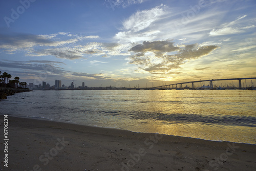 sand and palm trees line the shore of Coronado Island as the sun slowly rises above the Coronado Bridge