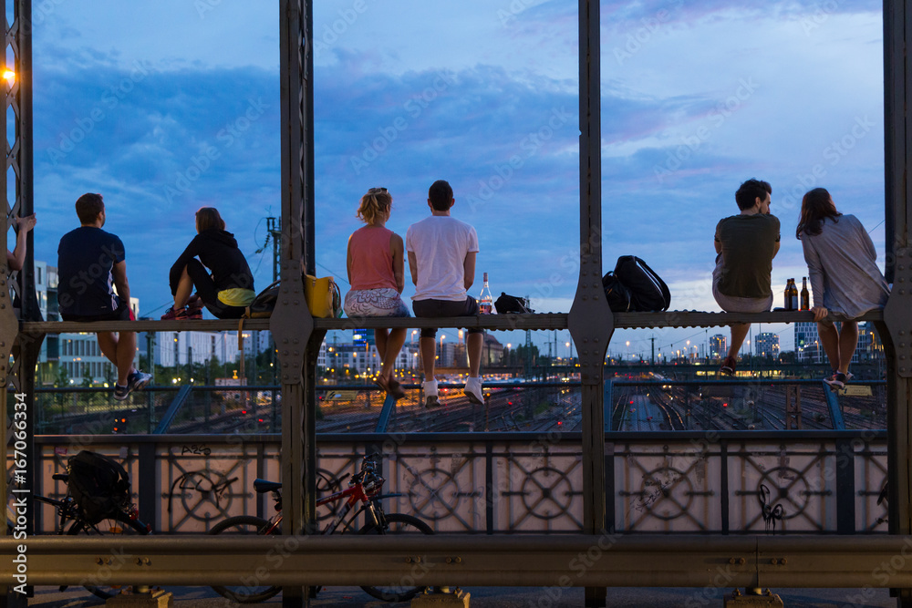 Young couples in love on romantic date sitting on railway bridge with a bottle of wine and watching the sunset over Munich urban city center. Just the two of us. - obrazy, fototapety, plakaty 
