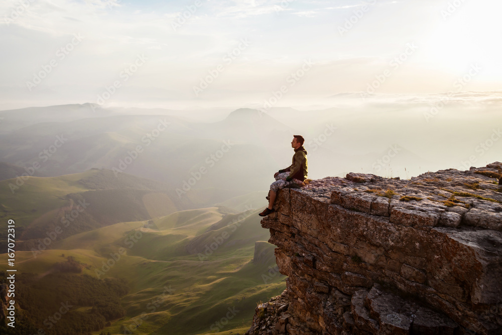 Man sitting on the cliff and enjoying sunset - obrazy, fototapety, plakaty 