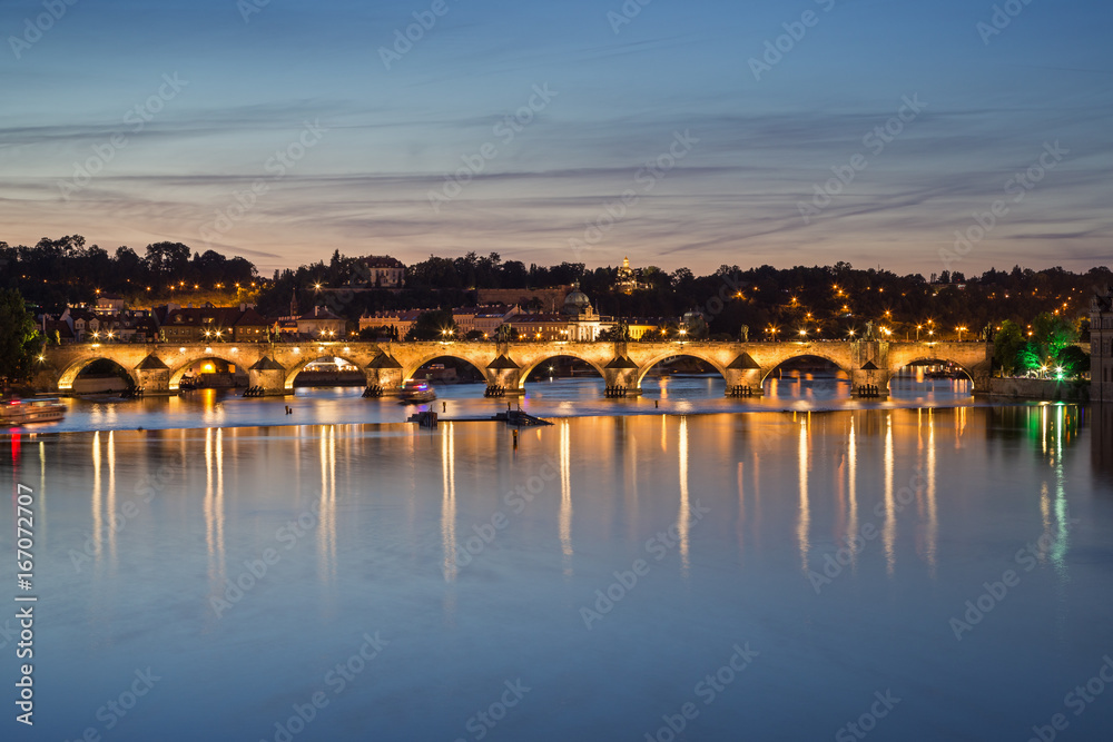 Lit Charles Bridge (Karluv most) and its reflections on the Vltava River in Prague, Czech Republic, at dusk.