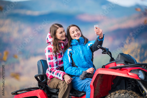 Close-up two happy girls in winter jackets on red ATV smiling and makes selfie on the phone with blurred background nature