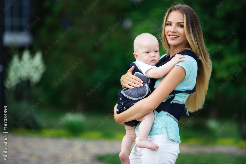 Fototapeta premium Pretty mother with baby boy in sling walking in green park