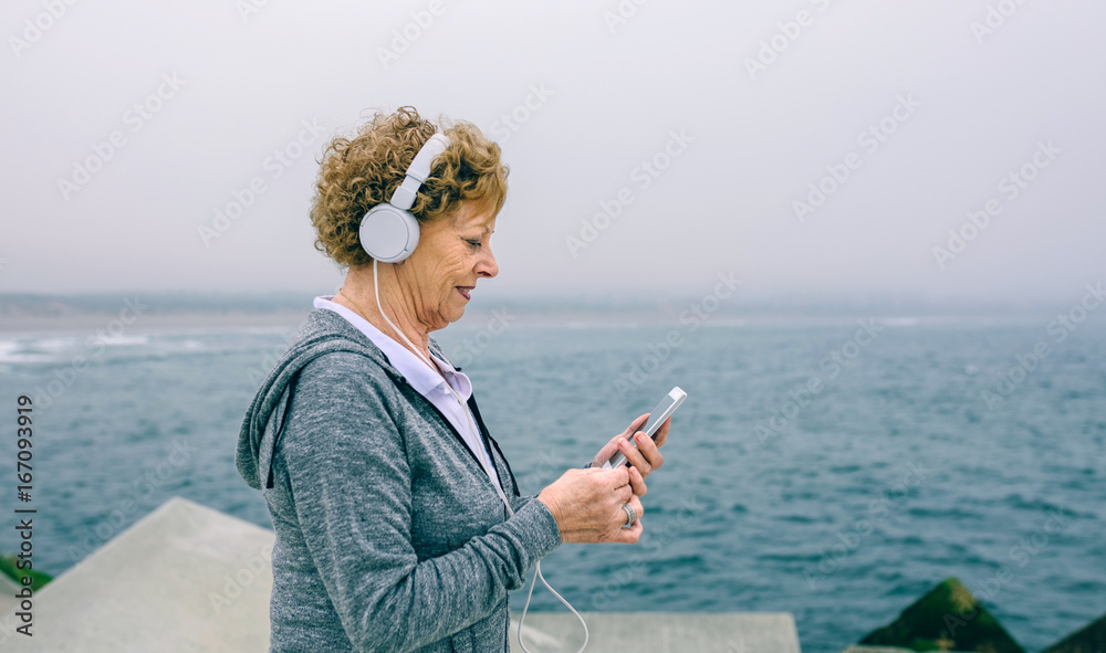 Senior sportswoman with headphones using her smartphone by sea pier