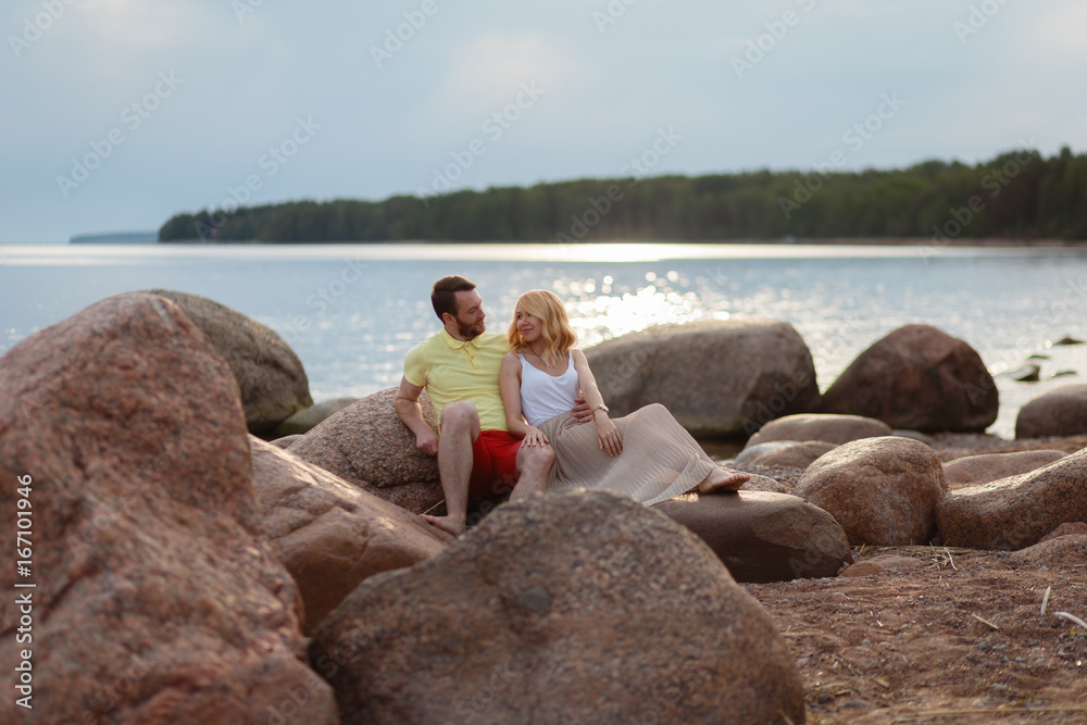 A man in shorts and a t-shirt and a girl in a summer dress enjoying each other. Beautiful couple sitting on a rock on the beach, watching the sunset. They wear casual clothes