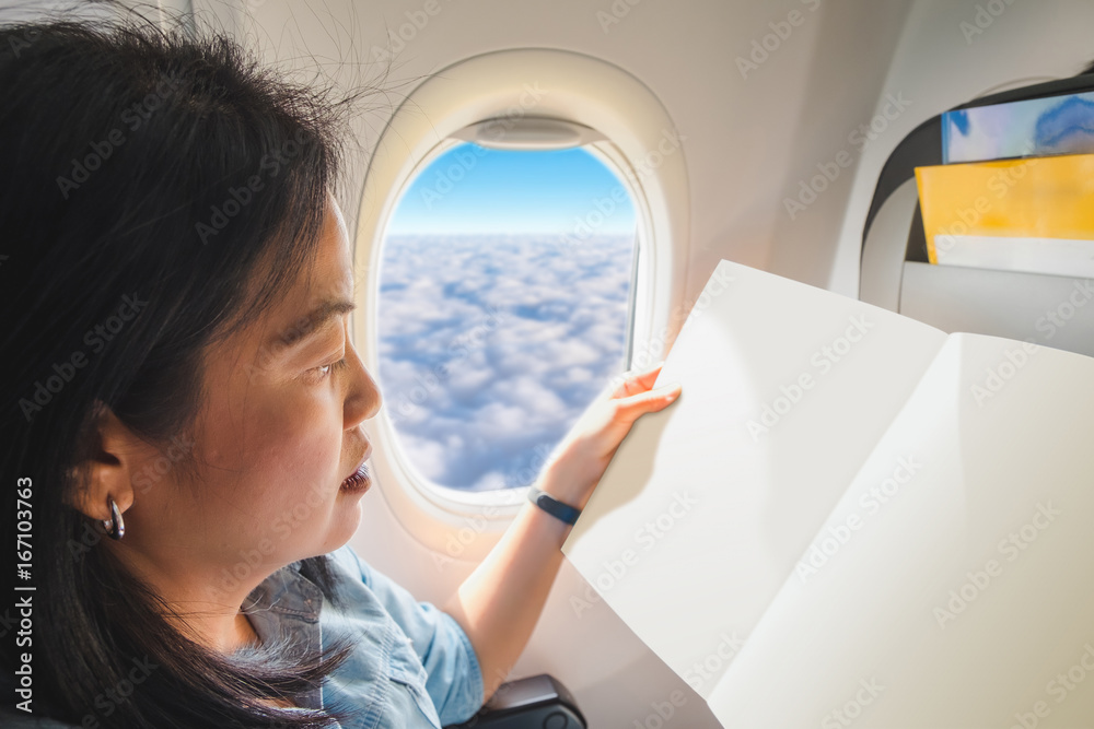 Obraz premium Asian Woman sitting at window seat in airplane and reading at blank open leaflet when flying,mock up for adding your design