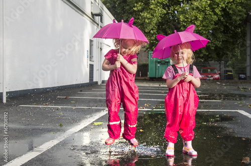 Children run through puddles on the street after a rain with pink umbrellas in hands, in pink bright rubber boots. The rays of the sun are reflected in the drops of water