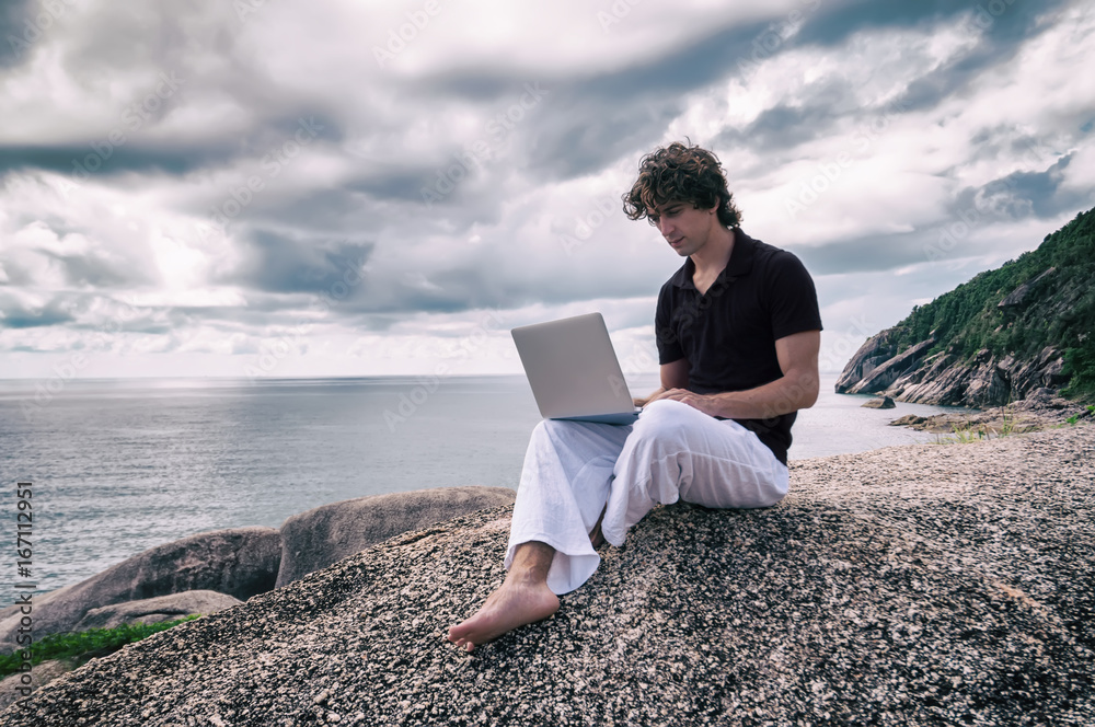 Young man, the freelancer sits on a high cliff over the sea and works remotely